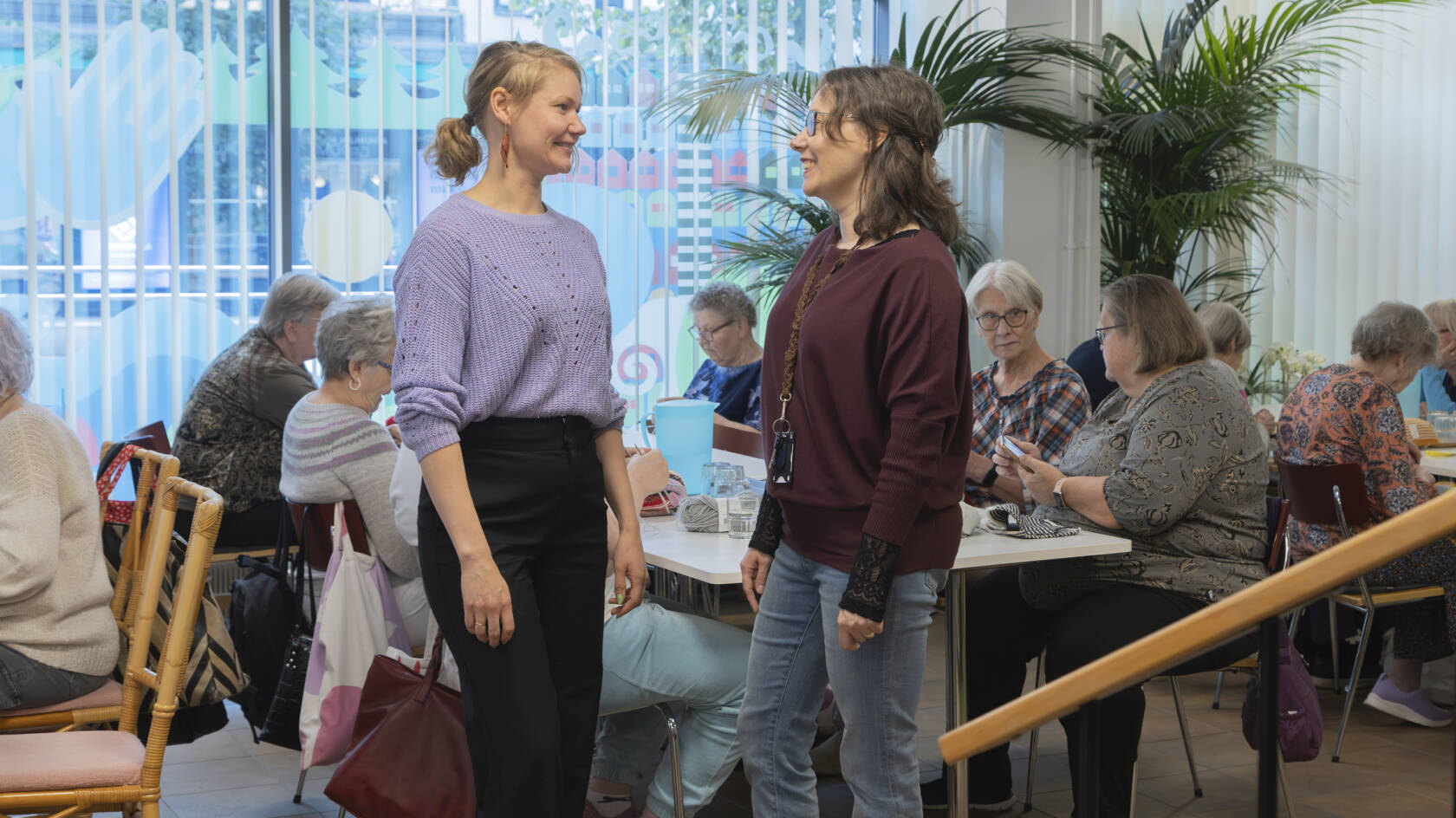 Kaksi naista keskustelee tilassa, jossa on taustalla iso ikkuna ja sen edessä ihmisiä toimimassa pöytien ääressä
