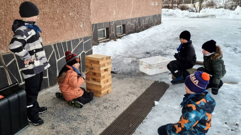 Kolme poikaa on polvillaan lumisessa maassa, yksi seisoo rakennuksen seinustalla ja yksi tarkastelee puupalikoista koottua rakennelmaa.