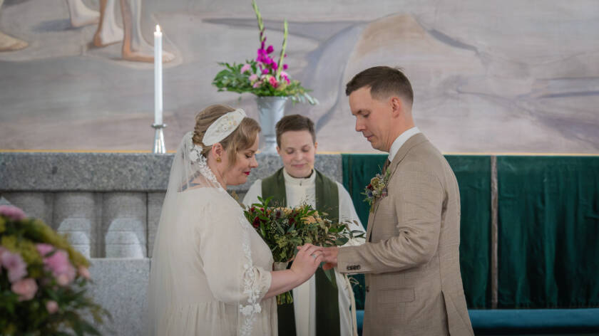 Nainen ja mies vaihtavat sormuksia alttarilla. Pappi hymyilee taustalla.
